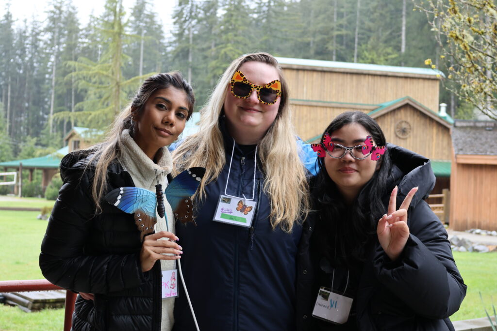 Three friends smiling outdoors with colorful butterfly accessories and casual jackets in nature.
